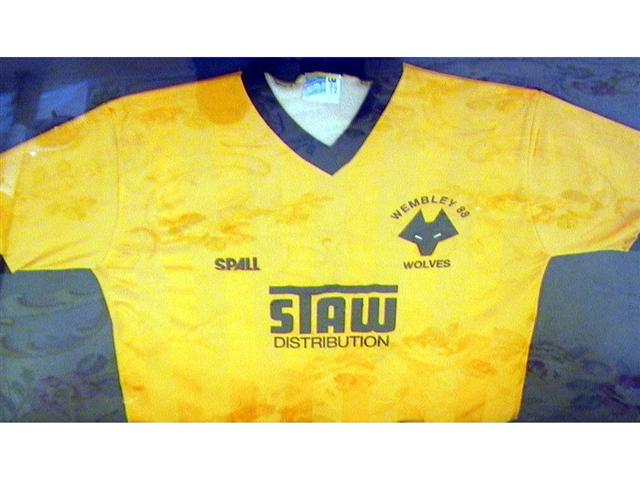 459. Shirt: An old gold with black trim V-neck shirt, Wolverhampton Wanderers crest to front with &lsquo;Wembley 88&rsquo; above and sponsors &lsquo;Staw Distribution&rsquo; below. Mounted, framed and glazed. Illus. &pound;500-600